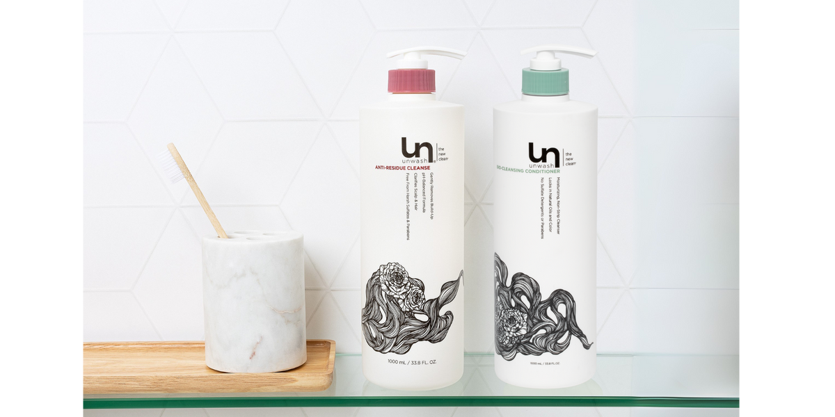 Unwash liter-size shampoo and cleansing conditioner bottles on a glass shelf in a modern bathroom, designed for hydration, balance, and better haircare value.