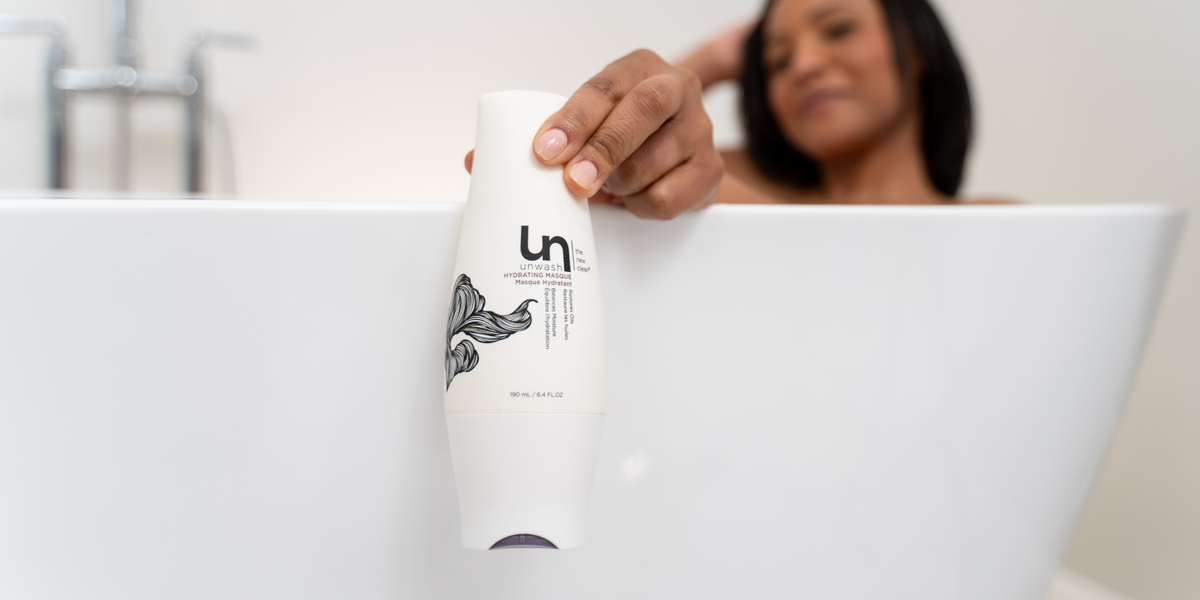 A woman relaxing in a bathtub reaches for a bottle of Unwash Hydrating Masque, highlighting the product's sleek white packaging with black floral design.