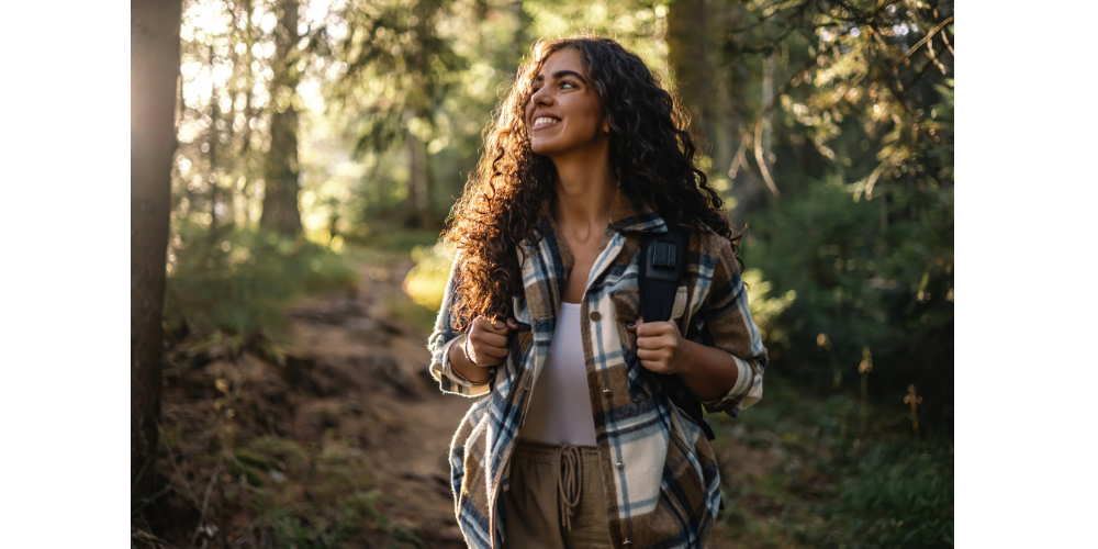 A woman with long, curly hair smiles confidently while hiking through a sunlit forest, wearing a plaid jacket and backpack, embracing her natural texture.